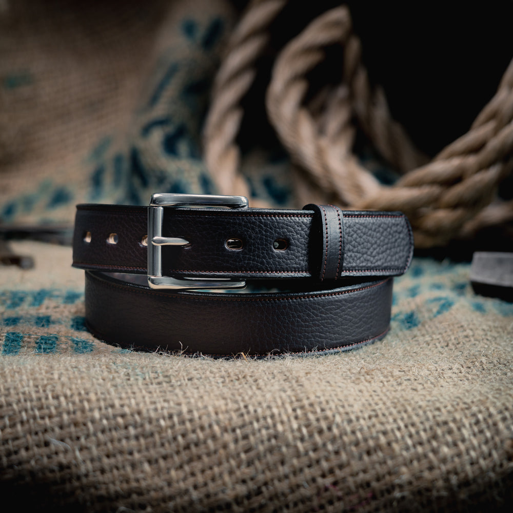 The Tuscan Embossed Lined by Hanks Belts, crafted from English Bridle Leather with a stainless steel buckle, is coiled on textured burlap with thick rope and a blurred background for a rustic touch.