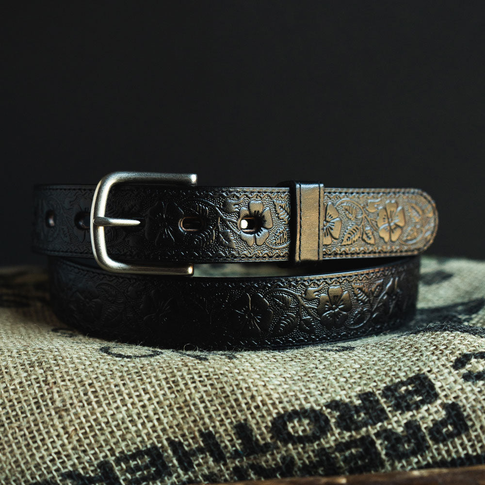 The Floral Embossed belt by Hanks Belts, crafted from full-grain black leather with a silver buckle and a floral embossed pattern, is coiled on textured burlap with partially visible black text.