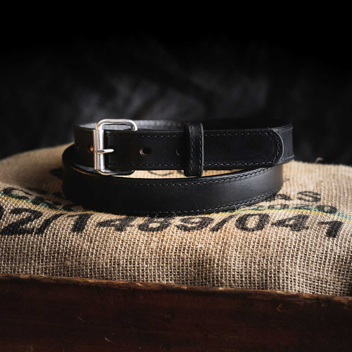 The Xtreme Belt - 1.25" by Hanks Belts, a durable leather gun belt with a heavy-duty roller buckle, is coiled and displayed on a burlap sack. The dark background creates a striking contrast, highlighting the belt's texture and intricate details.